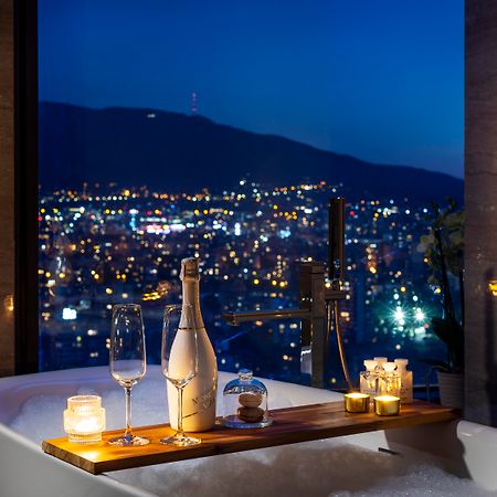 Grand Hotel Millennium Sofia Luaran gambar The photo features a luxurious bathtub placed by a large window, showcasing a scenic city view at night. There is a wooden tray resting on the edge of the tub, displaying a bottle of champagne, two glass flutes, and several lit candles, creating an a