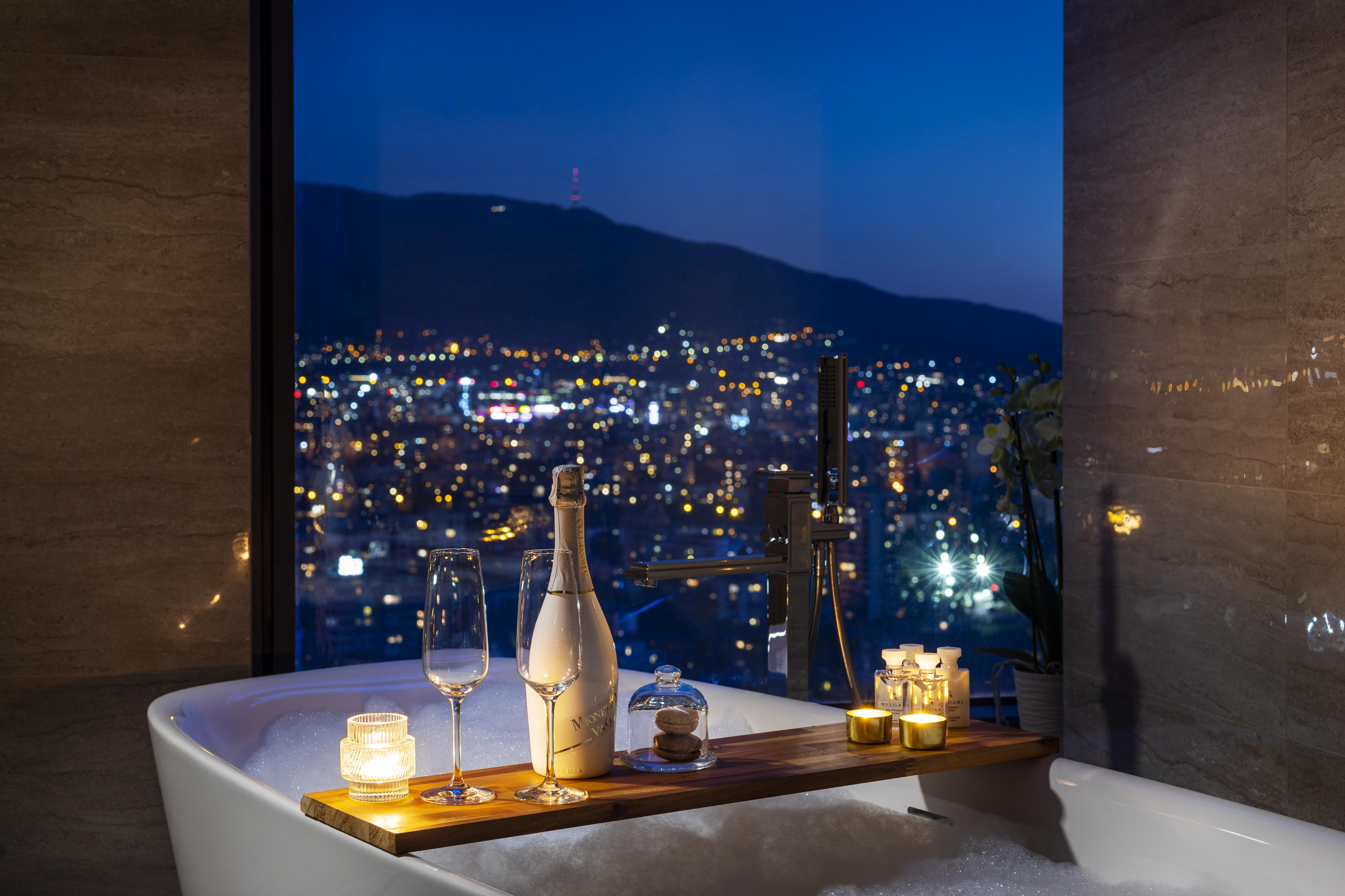 Grand Hotel Millennium Sofia Luaran gambar The photo features a luxurious bathtub placed by a large window, showcasing a scenic city view at night. There is a wooden tray resting on the edge of the tub, displaying a bottle of champagne, two glass flutes, and several lit candles, creating an a